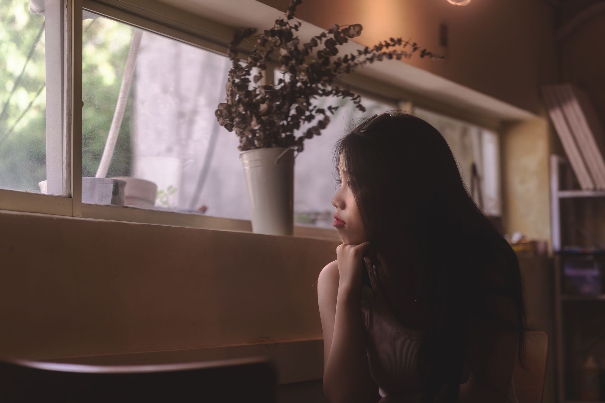 A woman gazes thoughtfully out of a window, surrounded by soft indoor lighting and decor.