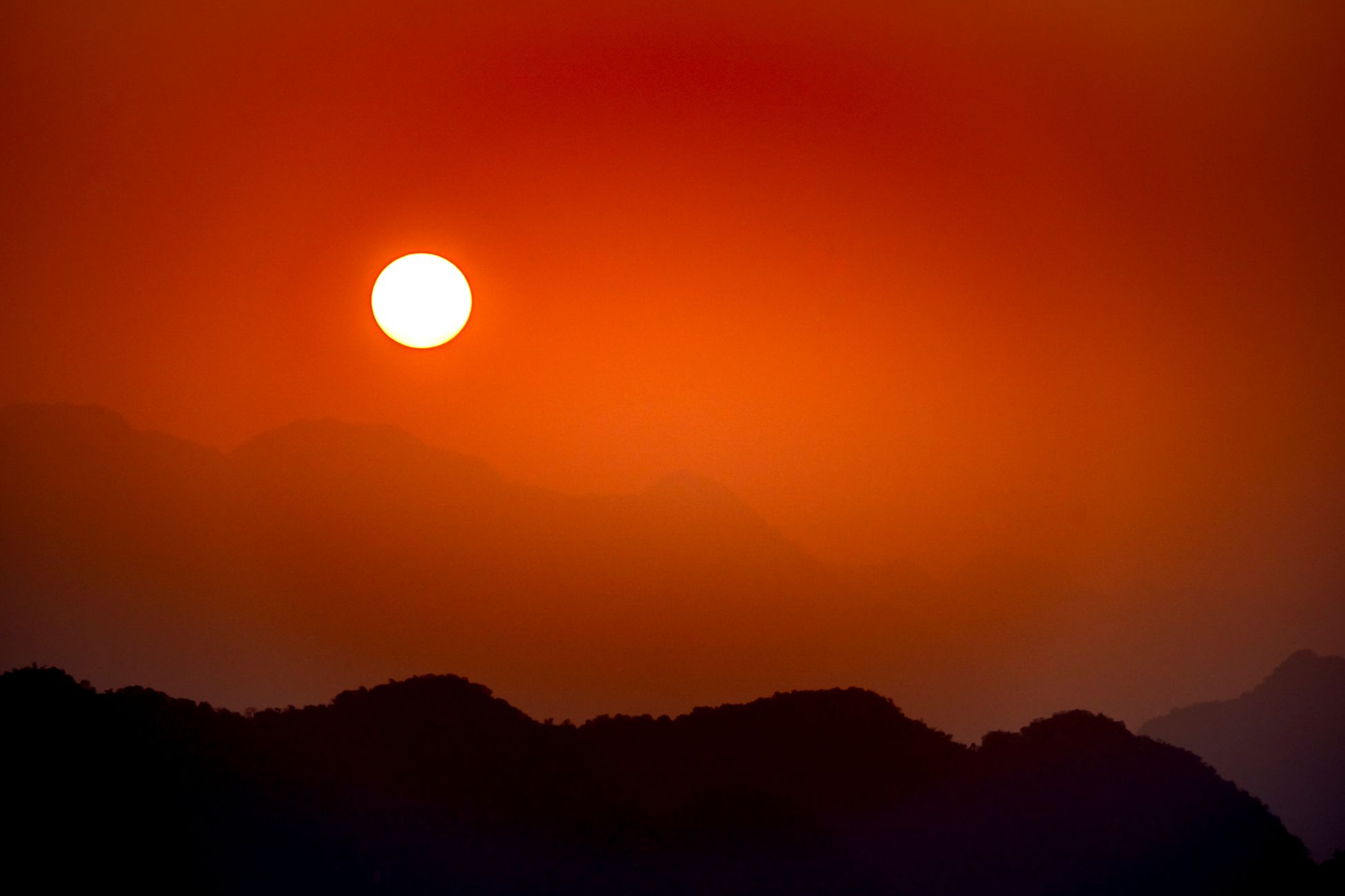 Bright sun shining on red cloudless sky over highland area with silhouettes of mountain ridge covered with mist at sundown time